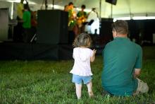 A toddler girl standing in the grass next to her sitting father as they watch an outdoor performance