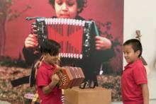 Two young boys play an accordion in front of a large picture of someone playing an accordion