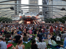 The silver Pritzker Pavilion in with audience in front sitting on grass