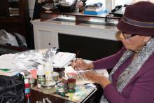 an older African American woman in a purple hat painting in colors on an art work in progress