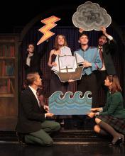 Six actors performing, holding cut outs of waves, a ship, clouds, and lightning