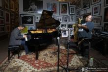 A man plays piano and another plays saxophone in a small art gallery