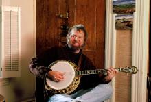 A man with brown hair and a beard sits in a sunlit livingroom with his tenor banjo. A Celtic cross hangs in the foreground.