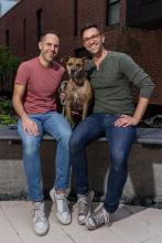 Two men sit on a stoop with a dog between them.