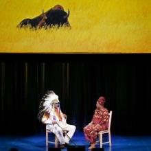 A man in Native American regalia sits on a stage across from a woman. An image of a buffalo is on the screen behind them.
