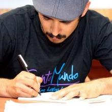 Headshot of a young Latino man wearing a hat and a Canto Mundo t-shirt bent over and writing on a page