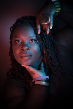 a close-up portrait of Ebony Noelle Golden, who is a dark-skinned Black woman with long locs. Her hands frame her face as she looks into the camera.
