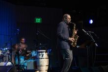 A man performs on the saxophone. Behind him is another musician performing on the drums