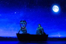 A stage production photo of two people ornately dressed in a boat with the moon behind them. 