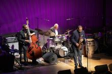 Three elderly Black men in suits playing drums, bass, and sax on stage with purple background. 