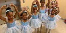 A group of young girls in leotards and tutus hold their hands above their heads in a ballet pose