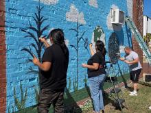 Three people stand in front of a brick wall painting a mural featuring an Earth and tall plants with a blue sky. 