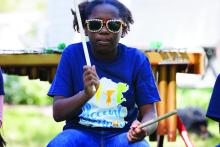Girl wearing sunglasses and blue shirt drumming on stage outdoors. 