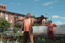 Black man with gray goatee wearing brown workshirt with arms extending in front of white woman in orange shirt doing same gesture, in a garden.