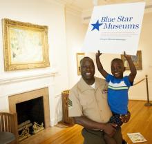 An African-American man in a Marine uniform holds his son in an art museum with artworks in the background