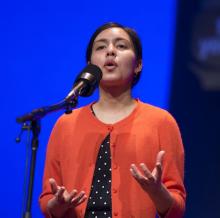 a young Hispanic woman wearing an unbuttoned orange sweater over a black dress with white polka dots recites into a microphone