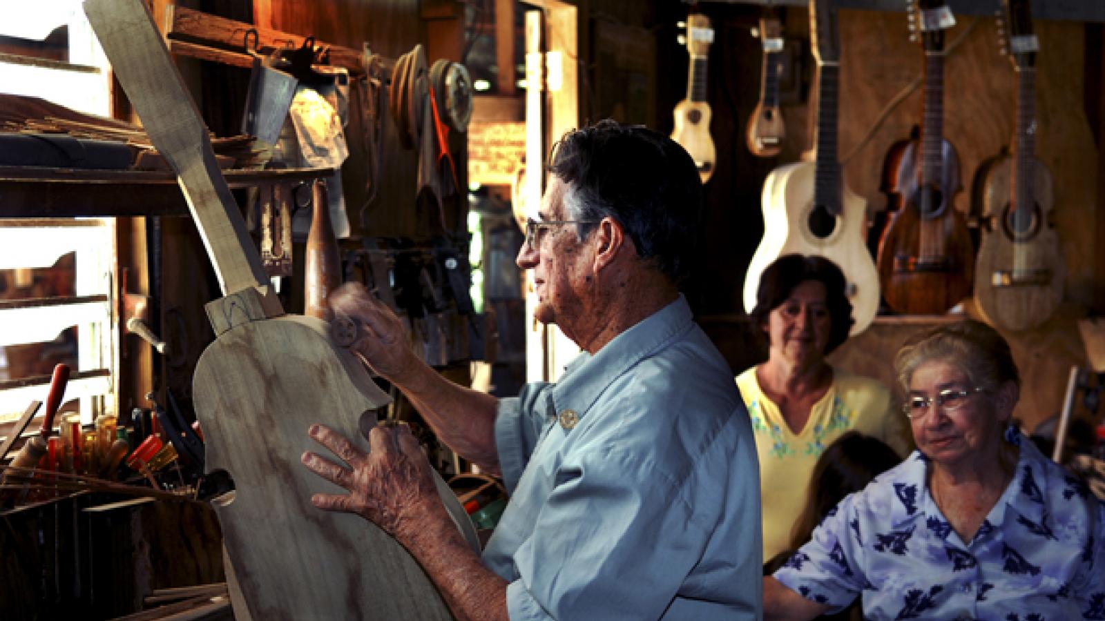A man works on building a stringed instrument with others behind him watching.