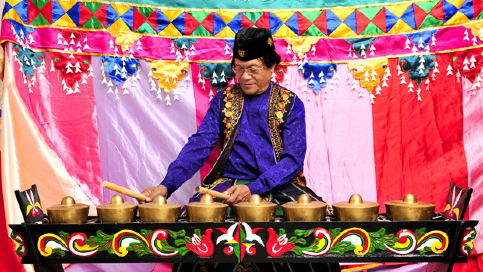 A man in a purple shirt and gold and black vest sits behind a musical instrument made up of eight small gongs.
