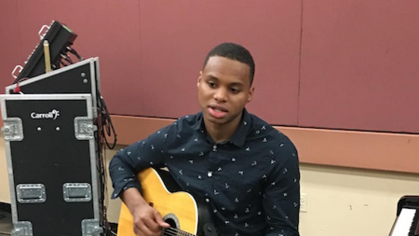 an African American young man sits on a piano stool holding his acoustic guitar