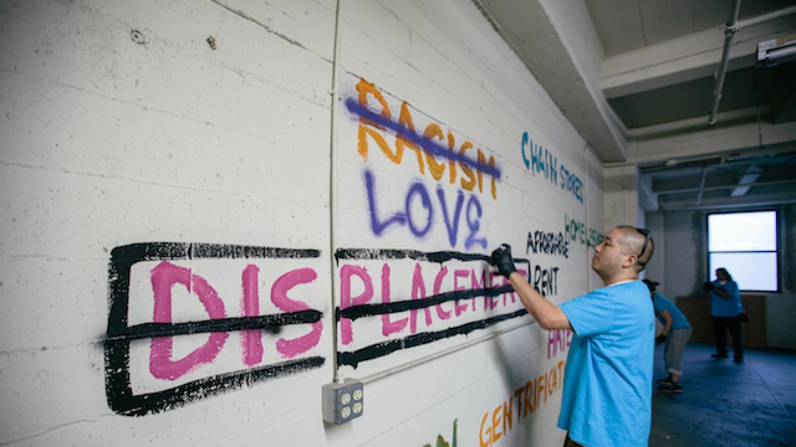 In a vacant space, artists spraypaint colorful text on a white cinderblock wall. Words include racism (crossed out), love, displacement crossed out, chain stores, homelessness, affordable rent, and gentrification.