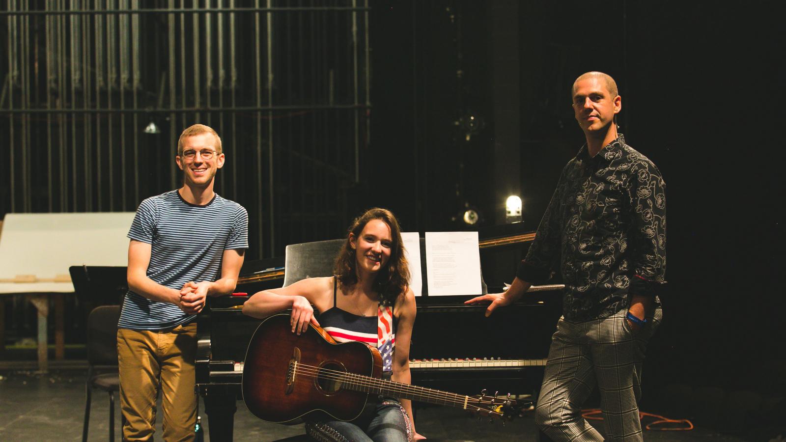 A young woman sits on a piano bench holding a guitar while two men stand casually leaning on the piano