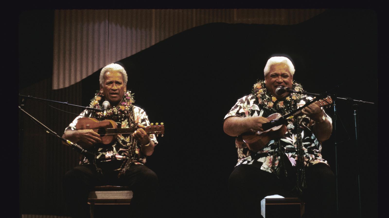 Two men on a stage playing ukuleles and singing.