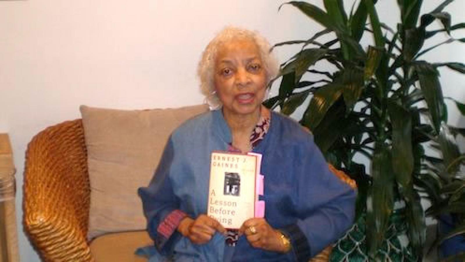 Ruby Dee on her couch holding up a copy of Ernest Gaines A Lesson Before Dying
