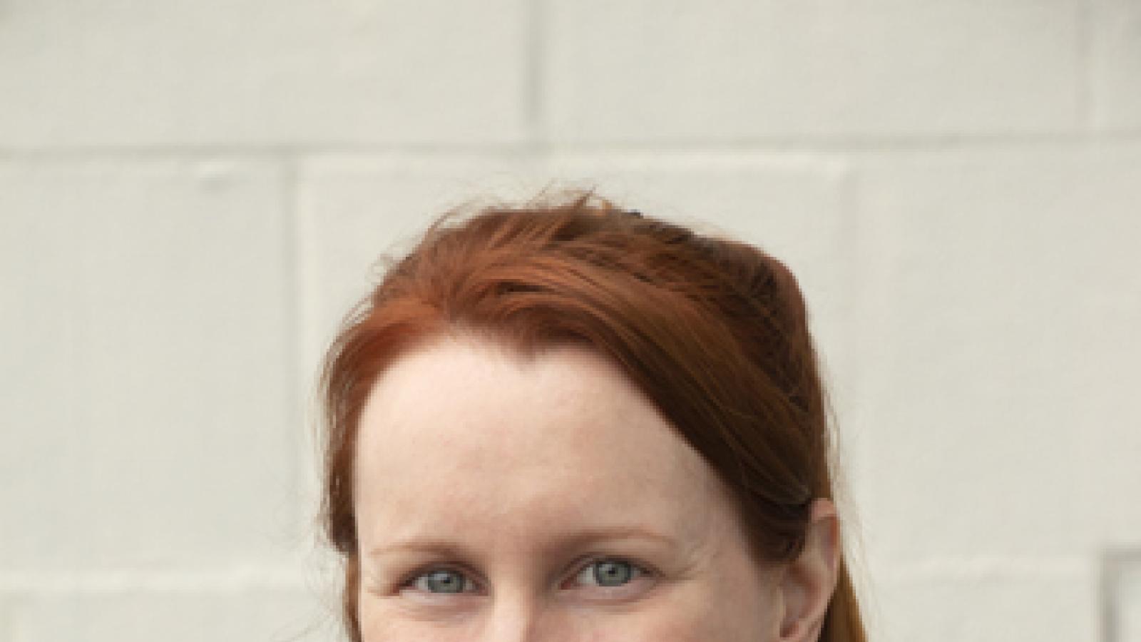 A woman with red hair stands against a white wall.
