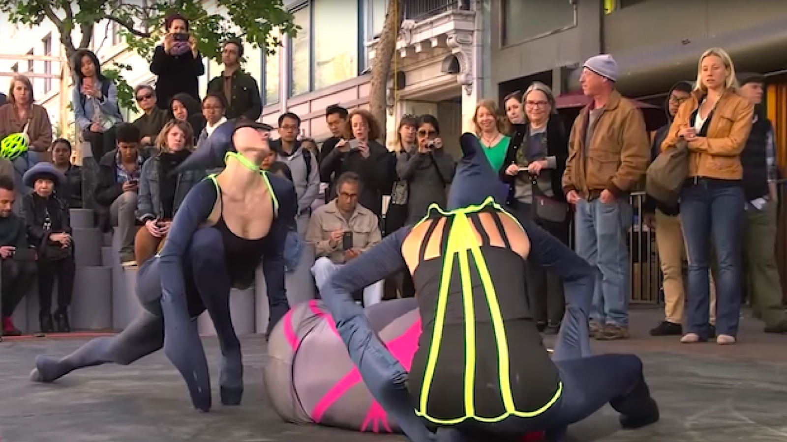 Dancers dressed as bugs dance on a city sidewalk for a crowd.