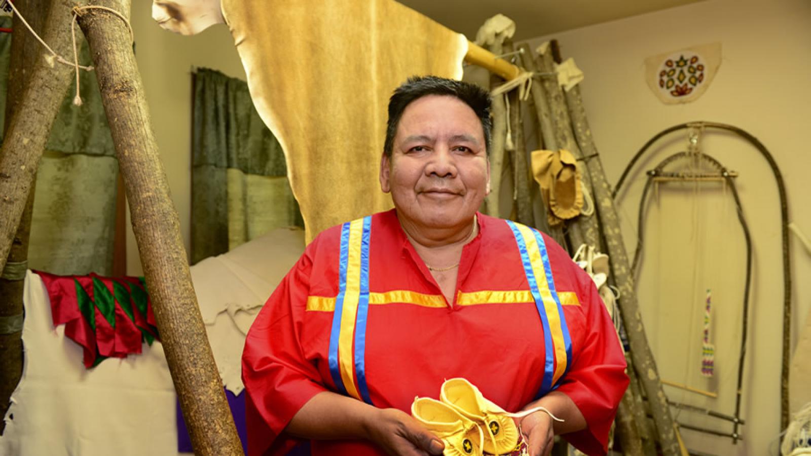 Standing in front of some of the tools of his craft, Gerald Hawpetoss smiles and holds a pair of small moccassins and an example of his beadwork.