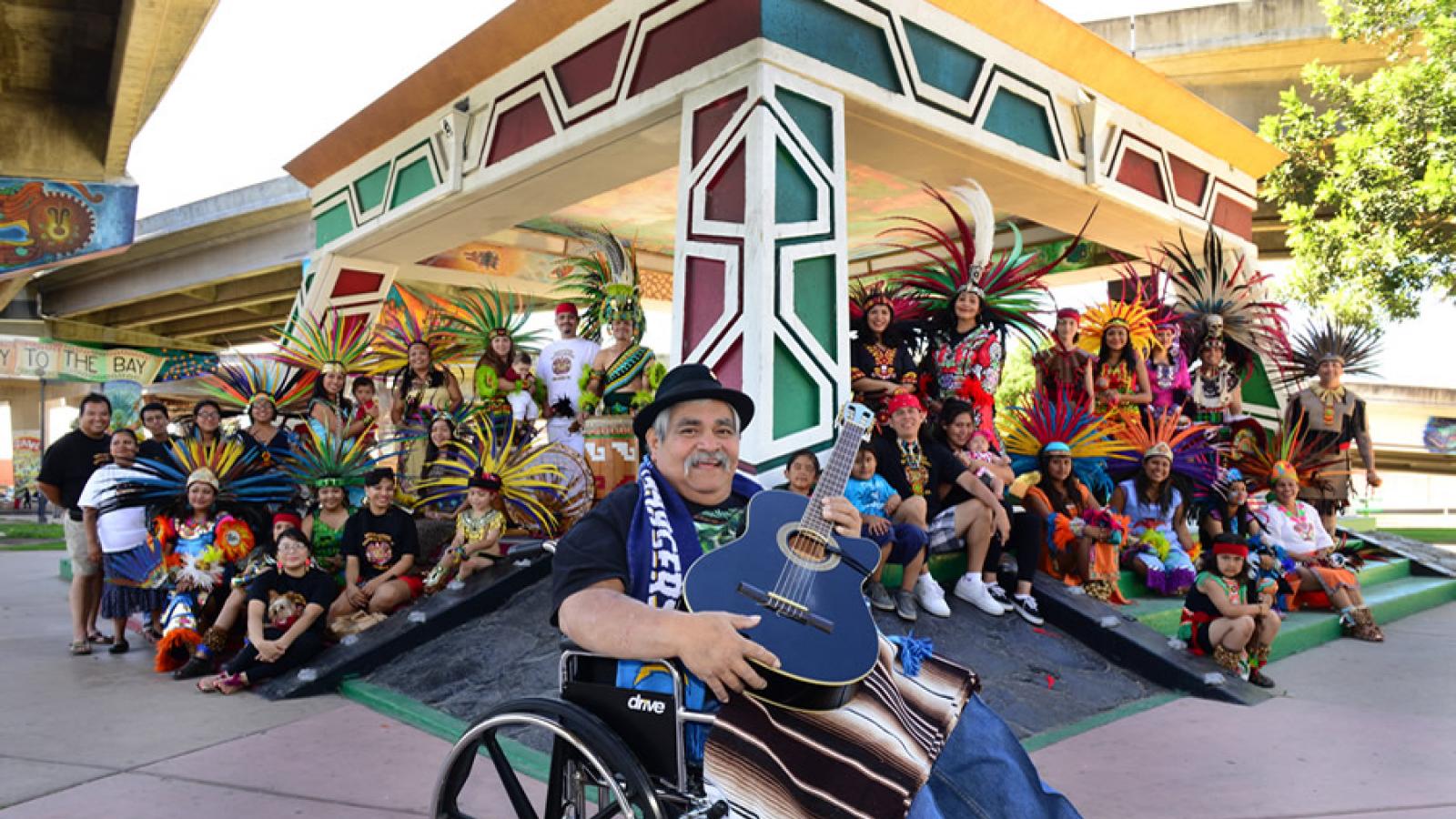 Ramón "Chunky" Sánchez sits in a wheelchair, holding a guitar and smiling, in front of a large group of communitiy members and performers wearing colorful and elaborate Aztec costumes.