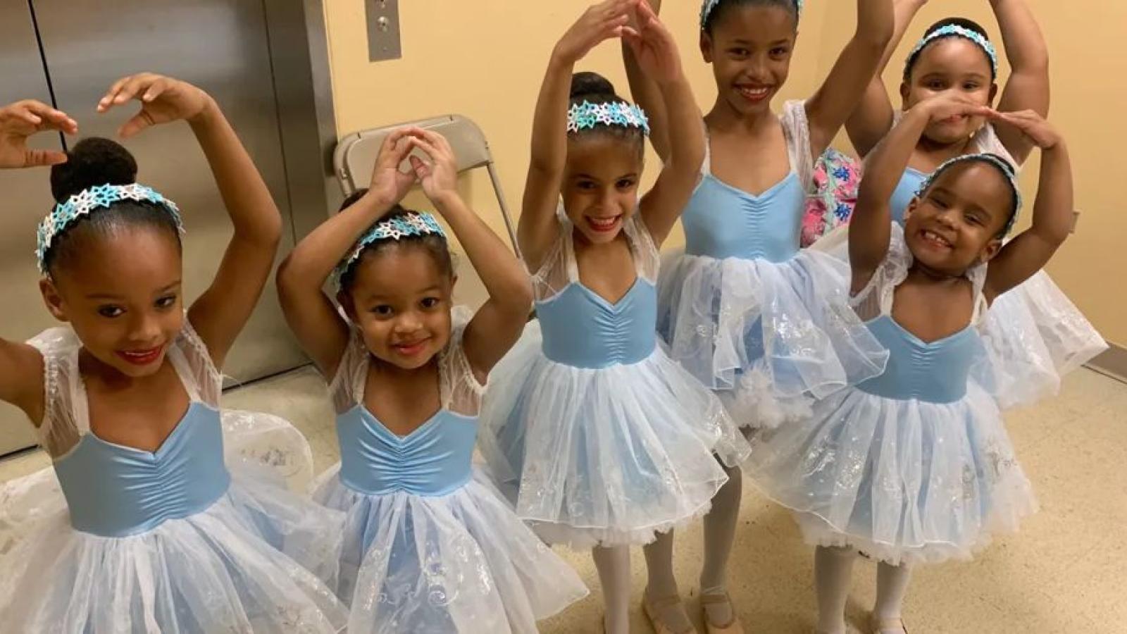 A group of young girls in leotards and tutus hold their hands above their heads in a ballet pose