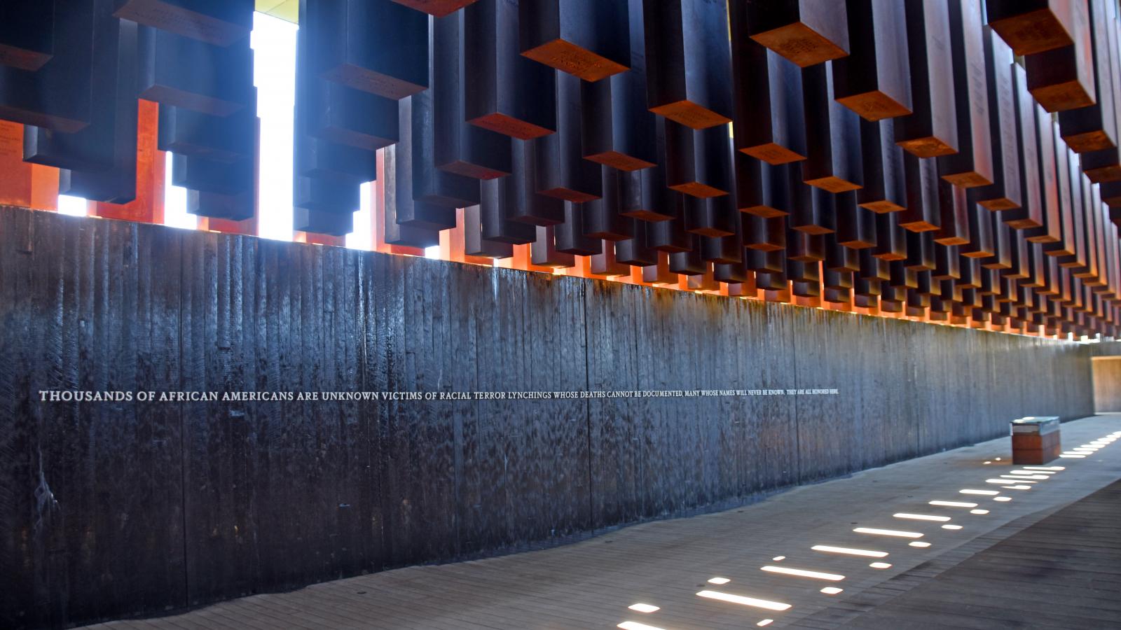 Large metal columns hang suspended in the air next to a stone wall as part of the National Memorial for Peace and Justice