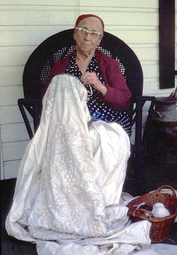 A woman sitting on chair working on a notted bed spread.