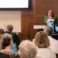  A group of people are seated in a room looking at four people seated at a table facing them and another woman behind a podium presenting a slideshow. 