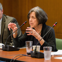  A man and two women sit behind a table at the front of a room. The woman in the middle is speaking. 