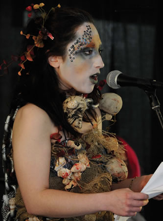 A heavily made-up performer wearing a costume of sea shells and sisal stands at a microphone and reads from a script