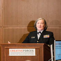  Two women are seated behind a table at the front of a room. A woman in a military outfit stands behind the podium at the front of the toom speaking. 
