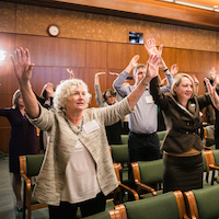  A group of men and women in the audience stand smiling with their hands raised up. 