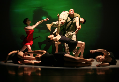 Man in military uniform holding another man in military uniform over his shoulders with a woman in black bent in front of them, a woman in red to their right reaching toward them, and three woman on the ground making a circle in front of them.