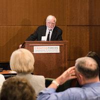  A man stands behind a podium at the front of the room looking out into the audience. 