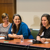  Four women sit behind a table at the front of the room smiling at each other. The woman on the right end is speaking. 