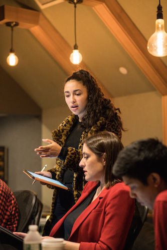 A young woman holding an iPad stands over other women working at a table