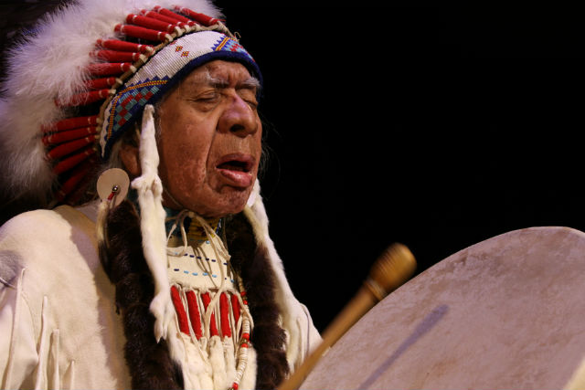 Man wearing Native headdress beats a handheld drum
