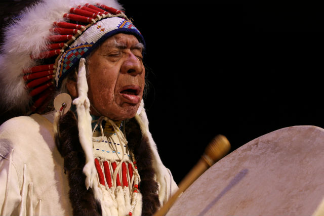 Man wearing Native headdress beats a handheld drum