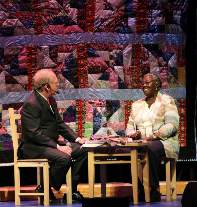 Woman sitting in front of hanging quilt she has made is interviewed by a man