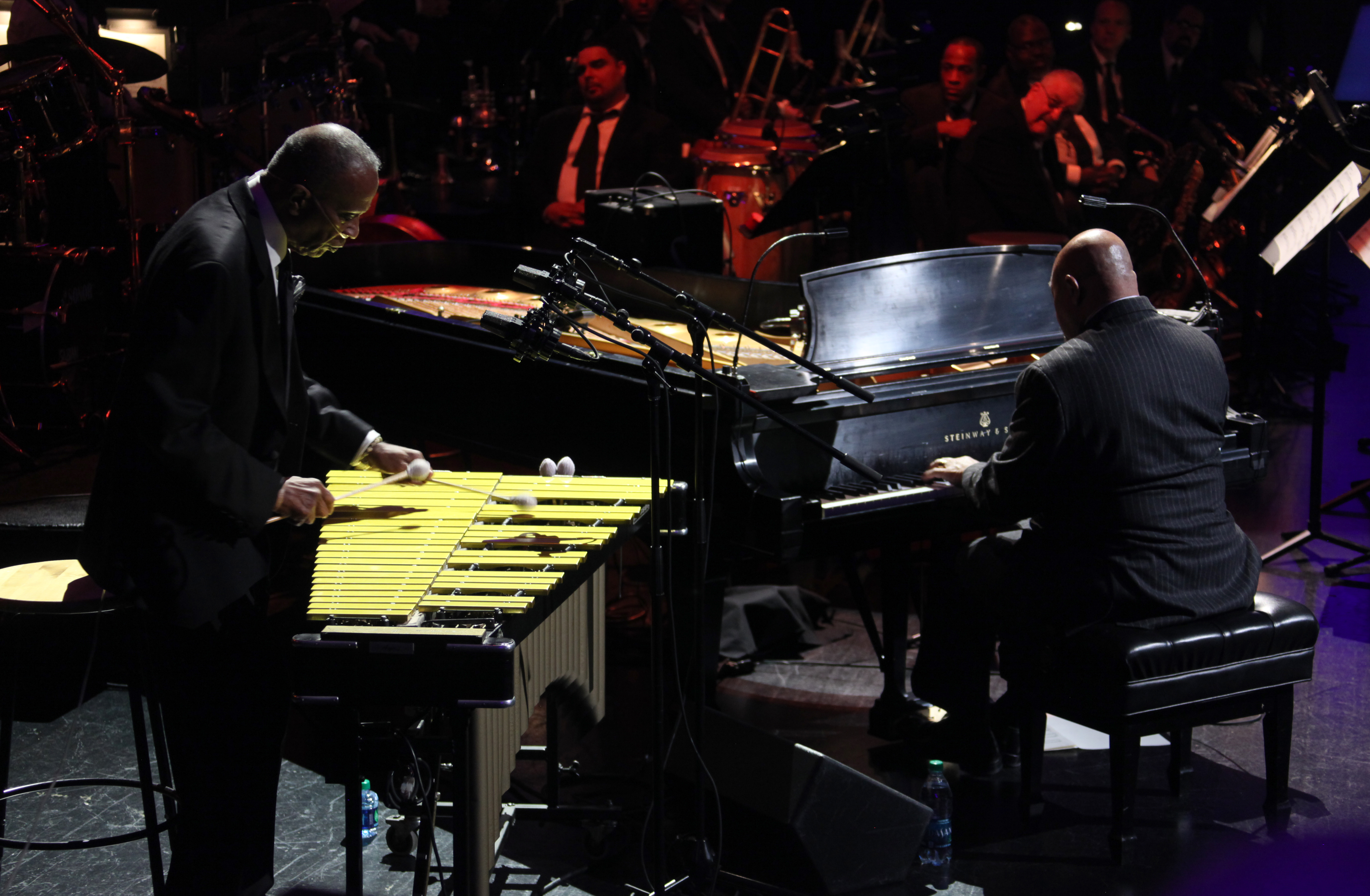 A man performs on the vibraphone next to another man performing on the piano.