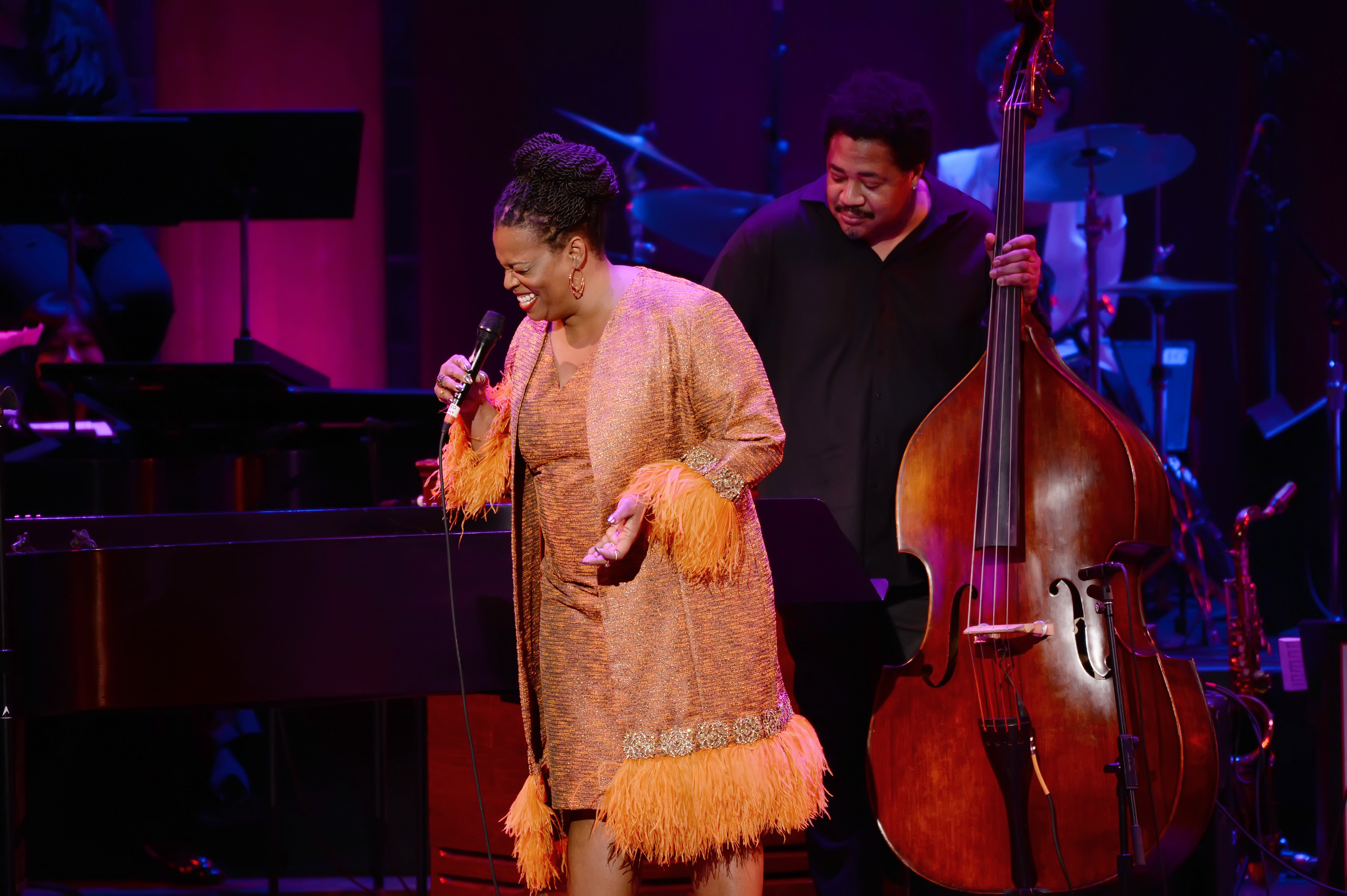 Woman in orange dress singing, with man holding a bass guitar behind her. 