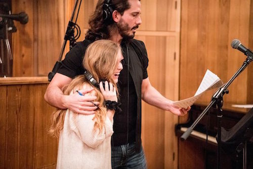 a man wearing headphones stands with his arm around a young woman in front of a microphone