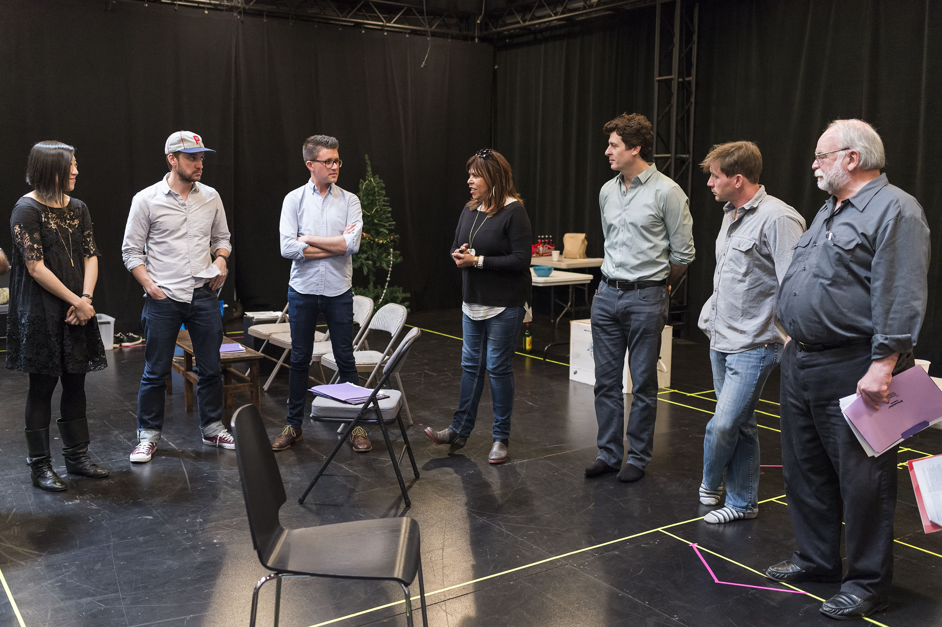 A group of actors, directors, and production staff standing in semi-circle on stage. 